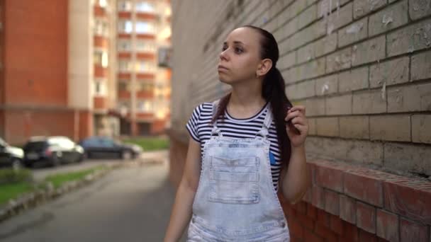 Młoda kobieta w luźnych ciuchach rozgląda się i przekręca włosy na ulicy. — Wideo stockowe