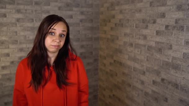 A beautiful emotional woman in a red hoodie is doing facepalm covering her face with palm. A young woman in a state of shock is covering her face with palm. — Stock Video