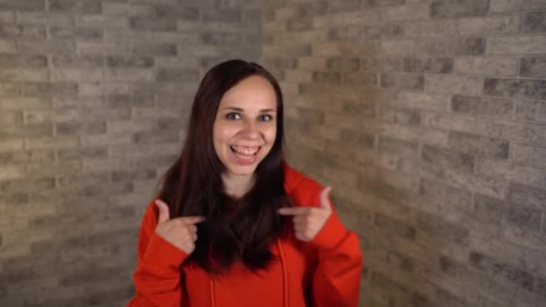 Een mooie jonge vrouw in een rode hoodie met lang haar glimlacht en toont haar handen op zichzelf op een grijze bakstenen achtergrond. — Stockvideo