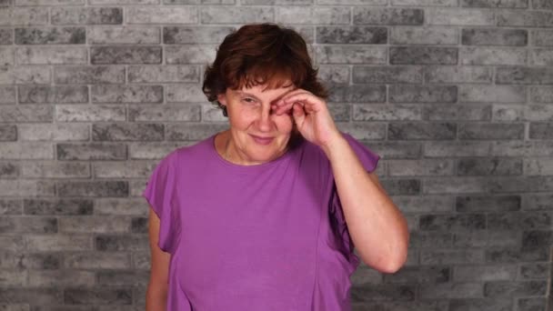 A beautiful mature woman in casual clothes with short hair smiles and rubs her eye on a gray brick background. — Stock Video