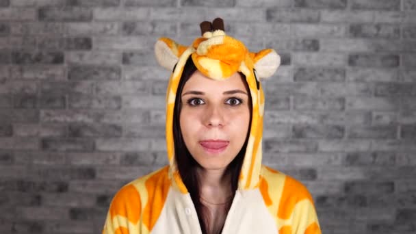 Uma mulher engraçada em um grande pijama de girafa lambe seus lábios e sorri em um fundo de tijolo cinza no estúdio. — Vídeo de Stock