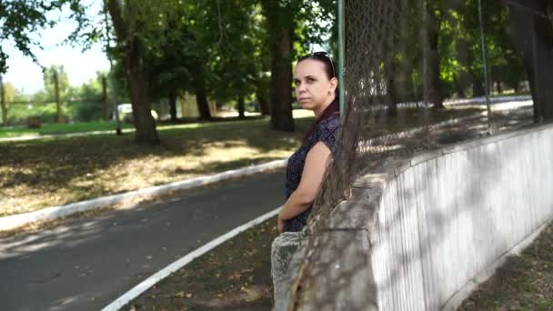Een mooie jonge vrouw in een sportpak kijkt weg en staat, leunend tegen het hek in het park. — Stockvideo