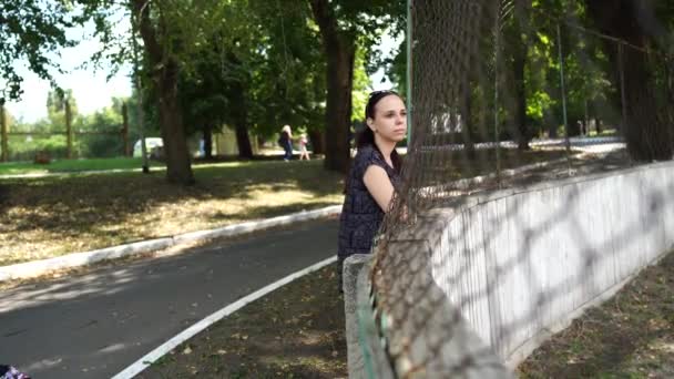Una joven guapa en traje deportivo mira hacia atrás y se pone de pie, inclinándose contra la valla del lattice en el parque.. — Vídeo de stock