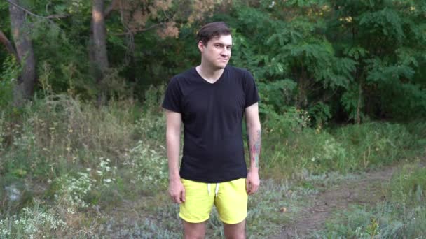 Ein junger gutaussehender Mann in lässiger Kleidung lächelt und zeigt im Wald mit dem Finger in die Kamera. — Stockvideo
