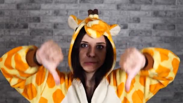 O femeie tânără frumoasă într-o pijama mare de girafă arată degetele în jos. O femeie arată un gest de dezaprobare pe un fundal de cărămidă. Conceptul de like sau dislike . — Videoclip de stoc