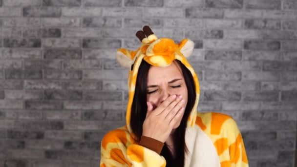 A beautiful young woman in a big pajamas of giraffe is yawning from fatigue on a gray brick background in the studio. — Stock Video