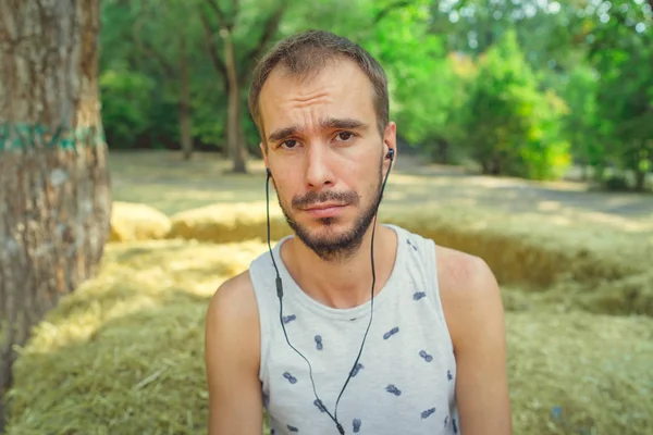 Un joven guapo con barba en una camiseta se sienta en el heno, escucha música y canta en el parque.. — Foto de Stock