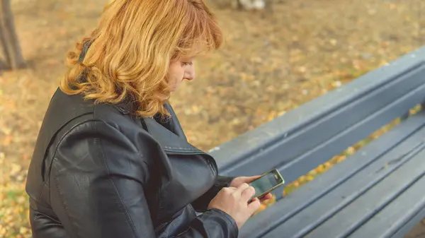 Eine erwachsene Frau, die im Herbst auf einer Bank sitzt, wählt mit dem Handy die Nummer — Stockfoto