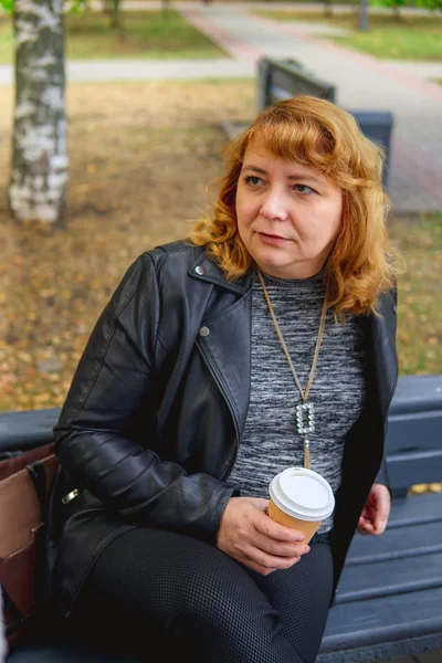 Volwassen vrouw die koffie drinkt in het najaarspark, zittend op een bankje — Stockfoto