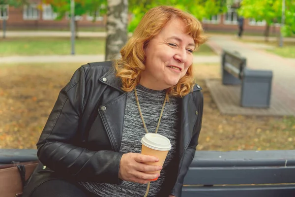 Volwassen vrouw die koffie drinkt in het najaarspark, zittend op een bankje — Stockfoto