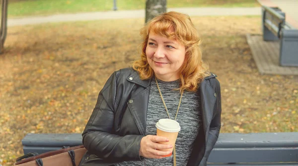 Volwassen vrouw die koffie drinkt in het najaarspark, zittend op een bankje — Stockfoto