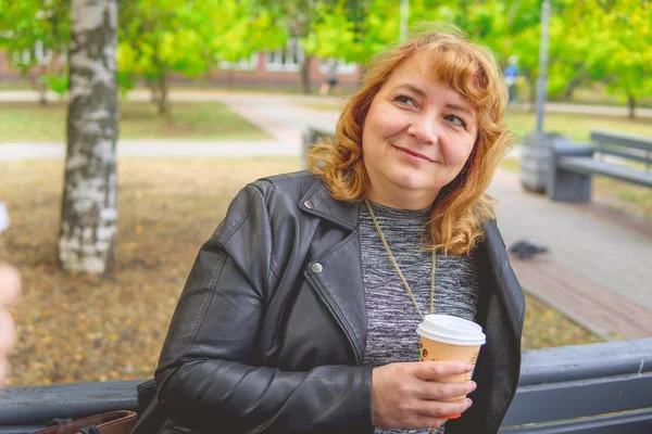 Volwassen vrouw die koffie drinkt in het najaarspark, zittend op een bankje — Stockfoto