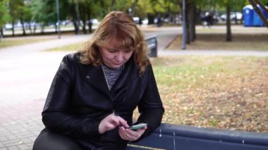 Sonbaharda bankta oturan ve cep telefonu kullanan yetişkin bir kadın..