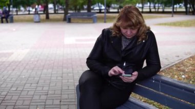 Sonbaharda bankta oturan ve cep telefonu kullanan yetişkin bir kadın..