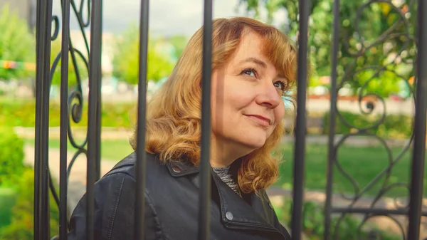 Mujer contra valla en el jardín. Mujer contenta que se ve enamorada mientras se encuentra en un parque verde contra la valla ornamental de hierro. — Foto de Stock