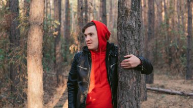 Güneşli bir günde ağaçların yanında duran adam. Ağaçların arasında poz veren gündelik kıyafetler giyen genç adamın portresi