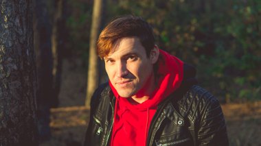 Güneşli bir günde ağaçların yanında duran adam. Ağaçların arasında poz veren gündelik kıyafetler giyen genç adamın portresi