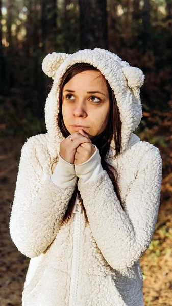 Egy fiatal nő fehér, bolyhos kapucnis pulcsiban összezsugorodik a hidegtől az erdőben.. — Stock Fotó
