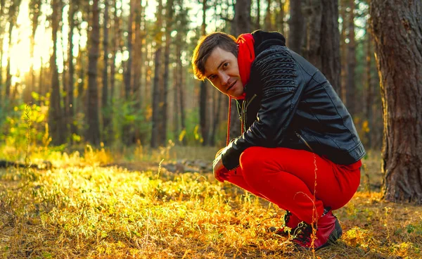 Un joven guapo con un traje rojo cálido y una chaqueta de cuero se sienta en sus cabellos en el bosque.. — Foto de Stock