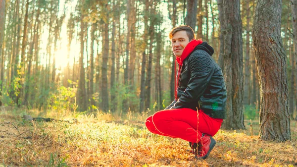 Un joven guapo con un traje rojo cálido y una chaqueta de cuero se sienta en sus cabellos en el bosque.. — Foto de Stock