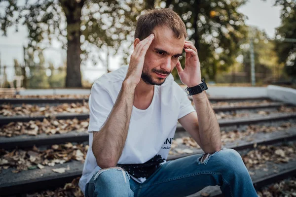 Hezký muž v depresi. Ne oholený hipster sedí na schodech v podzimním parku a drží si hlavu. Koncept: bolest hlavy a zármutek — Stock fotografie