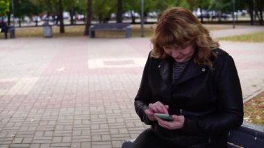 Sonbaharda bankta oturan yetişkin bir kadın Park cep telefonundaki numarayı çevirir.