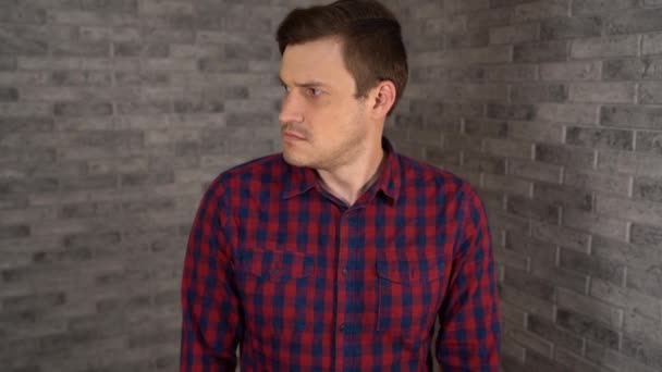 A young handsome man in plaid shirt looks around suspiciously on a gray brick background in the studio. — Stock Video