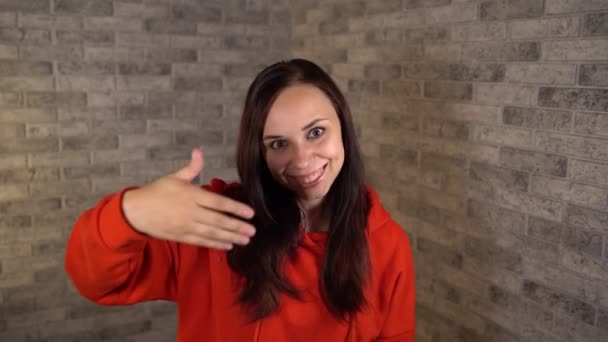 Una guapa jovencita en un hoodie rojo con largas sonrisas de pelo y olas de su mano, llamando a alguien en un fondo de ladrillo gris en el estudio.. — Vídeos de Stock