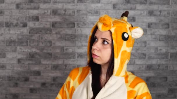 Красива молода жінка у великій піжамі жирафа підозріло виглядає на цегляному фоні в студії . — стокове відео