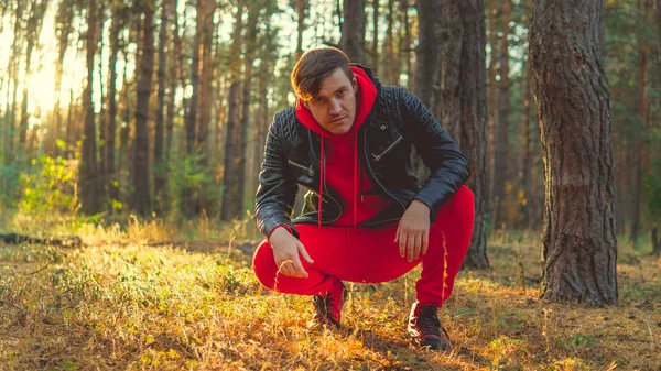 Un hombre guapo sentado en el bosque al amanecer. Un hombre con traje de tracksuit rojo y una chaqueta de cuero negro. — Foto de Stock