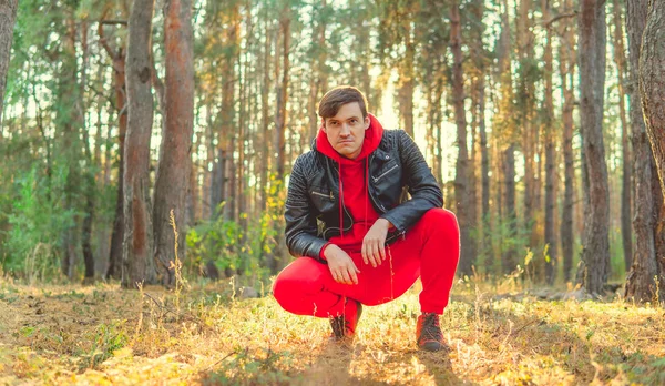 Beau homme assis en train de poser dans les bois au lever du soleil. Un homme vêtu d'une combinaison rouge et d'une veste de cuir noire — Photo