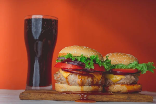 Zwei riesige saftige Hamburger stehen auf einem Schneidebrett und ein großes Glas mit Cola auf orangefarbenem Hintergrund.. — Stockfoto