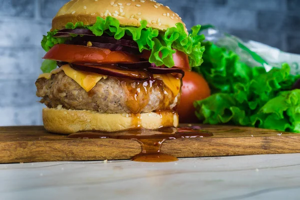 Um hambúrguer suculento enorme está em uma tábua de corte em um fundo de tijolo cinza . — Fotografia de Stock