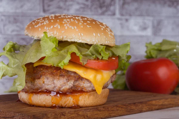 Obrovský šťavnatý hamburger je na sekání palubě se zeleninou na šedém cihlovém pozadí. — Stock fotografie