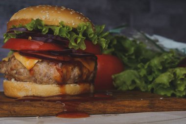 Kocaman sulu bir hamburger, gri tuğlalı arka planda sebzelerle doğrama tahtasının üzerinde..
