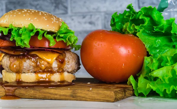 Um hambúrguer suculento enorme está em uma tábua de corte com legumes em um fundo de tijolo cinza . — Fotografia de Stock