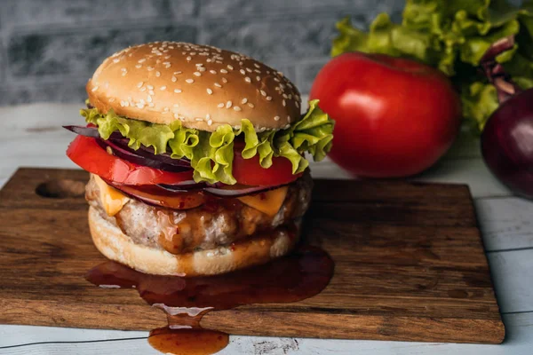 Um hambúrguer suculento enorme está em uma tábua de corte com legumes em um fundo de tijolo cinza . — Fotografia de Stock