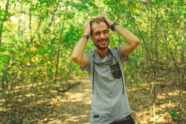 O cara está desfrutando de uma caminhada no Parque. Um homem experimenta êxtase, parado na floresta e segurando sua cabeça. Hipster sorri e desfruta — Fotografia de Stock