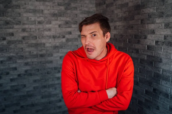 Um homem bonito novo em um hoodie vermelho com winks bristles com boca aberta em um fundo cinzento do tijolo. — Fotografia de Stock