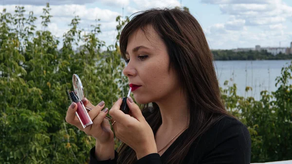 Una giovane bella donna dai lunghi capelli scuri dipinge le labbra di rossetto rosso, guardando in un piccolo specchio all'aperto. Avvicinamento. — Foto Stock