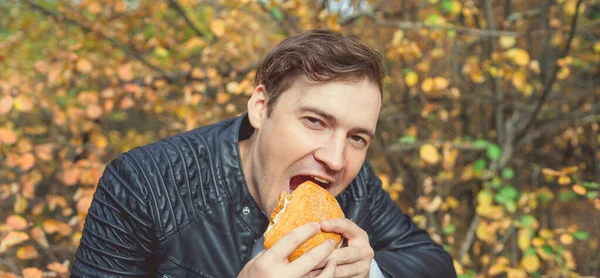 Een man die een hamburger eet in een zonnig lentepark. Jonge man met zonnige dag hamburger eten in de herfst bos. — Stockfoto