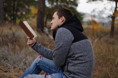 Güz ormanında kitap okuyan güzel bir adam. Bir adam sonbahar ormanında bir ağacın yanında oturuyor ve elinde bir kitap tutuyor. Erkek kitap okuyor. Sonbahar mevsimi konsepti ve okuma