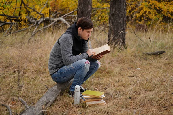 Gyönyörű férfi, aki könyvet olvas az őszi erdőben. Egy férfi ül egy fa mellett egy őszi erdőben, és könyvet tart. A férfi könyvet olvas. Őszi szezon koncepció és olvasás — Stock Fotó