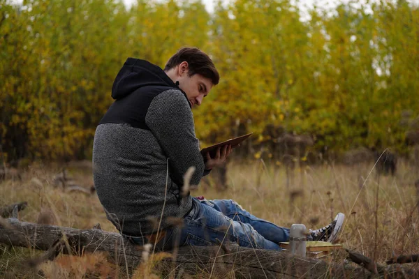 Hermoso hombre leyendo un libro en el bosque de otoño. Un hombre se sienta cerca de un árbol en un bosque de otoño y tiene un libro. Lectura masculina de un libro. Concepción y lectura de la temporada de otoño — Foto de Stock