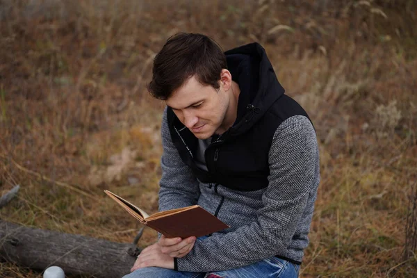 Gyönyörű férfi, aki könyvet olvas az őszi erdőben. Egy férfi ül egy fa mellett egy őszi erdőben, és könyvet tart. A férfi könyvet olvas. Őszi szezon koncepció és olvasás — Stock Fotó