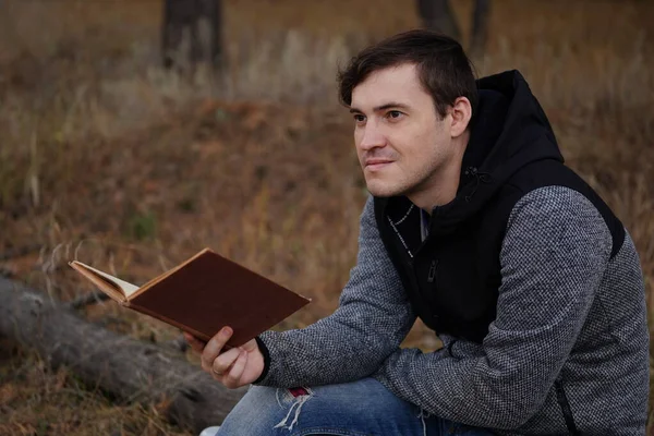 Güz ormanında kitap okuyan güzel bir adam. Bir adam sonbahar ormanında bir ağacın yanında oturuyor ve elinde bir kitap tutuyor. Erkek kitap okuyor. Sonbahar mevsimi konsepti ve okuma — Stok fotoğraf