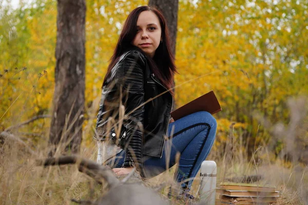 Młoda piękna kobieta z długimi ciemnymi włosami w luźnych ubraniach siedzi na kłodach i czyta książkę w jesiennym lesie.. — Zdjęcie stockowe
