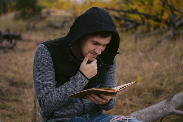 Een jonge knappe man in casual kleding zit op een boomstam en leest een boek in het herfstbos. — Stockfoto