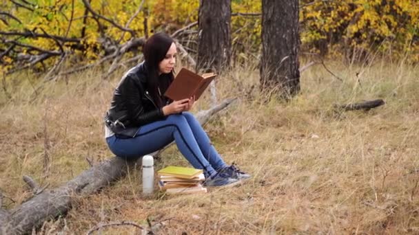 A young beautiful woman with long dark hair in casual clothes is sitting on a log and reading a book in the autumn forest. — Stock Video