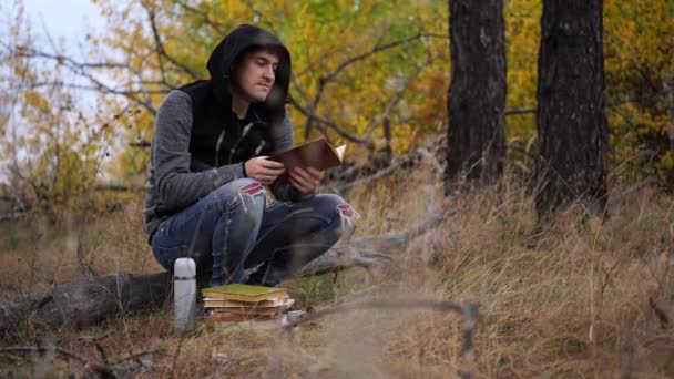 Um jovem bonito homem em roupas casuais está sentado em um log e ler um livro na floresta de outono. — Vídeo de Stock